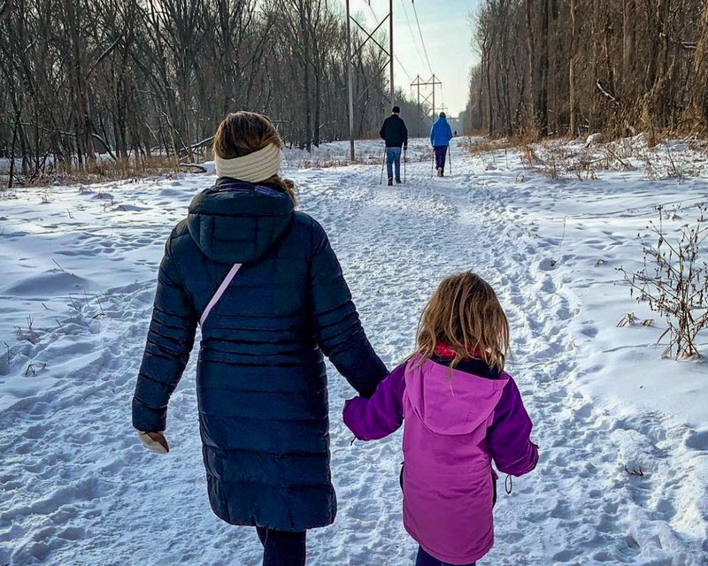 A mom and her young daughter walk across a snowy trail hand-in-hand, while exploring Fort Snelling State Park during the winter.