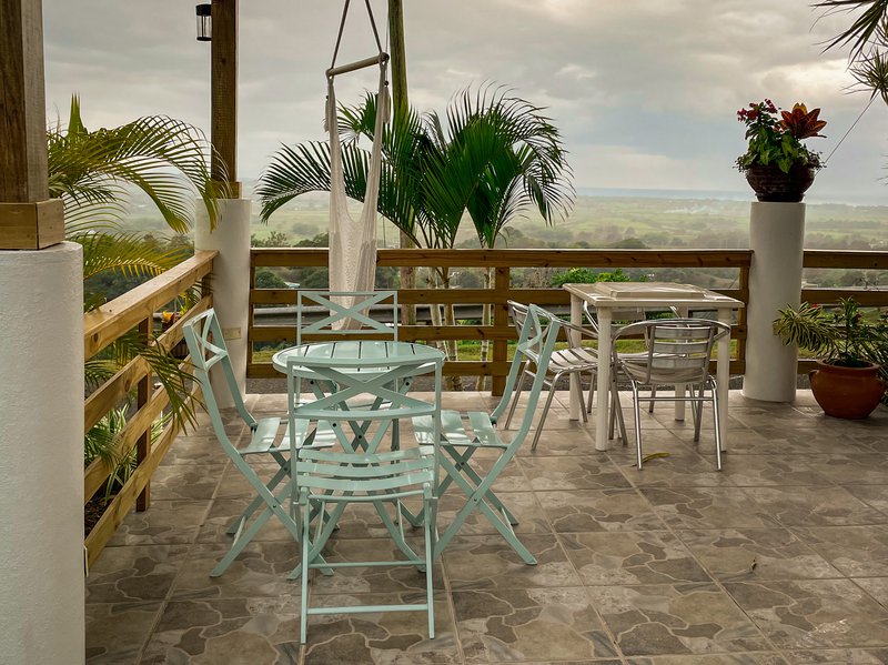 A patio featuring two tables with chairs, a hammock, lush greens, and a mountainous view from a Vrbo in Puerto Rico.