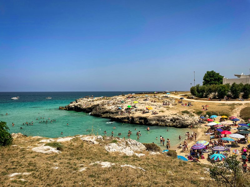 An aerial view of a beach in Monopoli, with turquoise waters and colorful umbrellas dotting the sand, while people enjoy the water and beach on a sunny day.