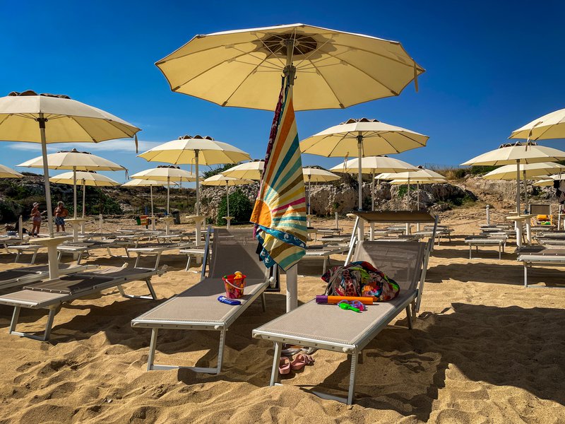 Towels hang from a lido umbrella, with two loungers resting underneath, protected from the sun on a beach in Puglia.