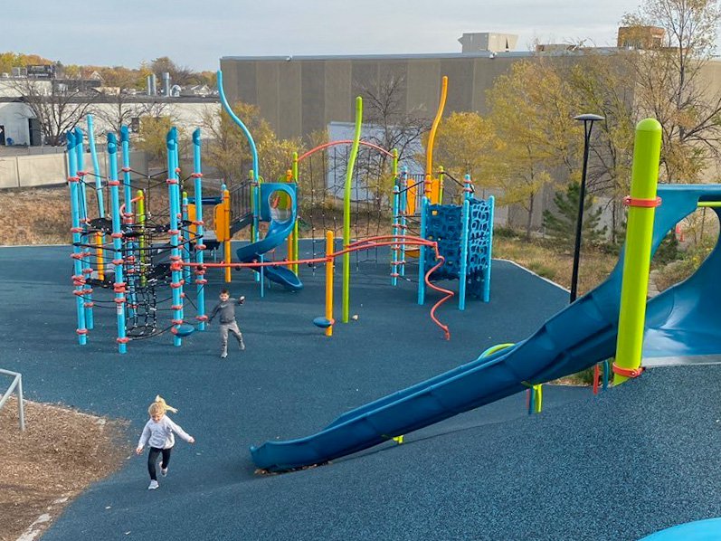 Two kids run around the urban playground, Midway Peace Park, featuring large slides and colorful equipment.